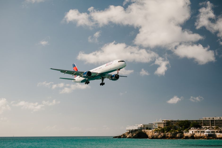 Commercial aircraft landing above ocean with clear blue skies, near beach resort.